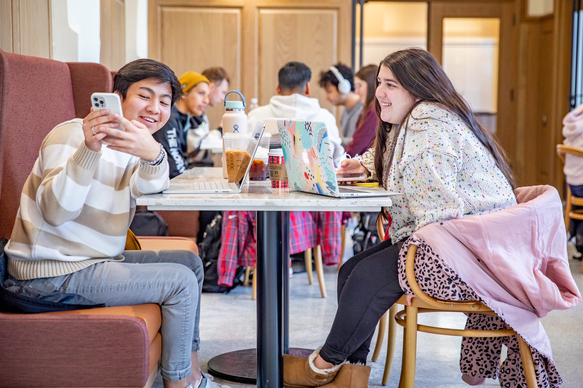 Two students sit at a table. One student is wearing a striped sweater and jeans and the other is wearing a cardigan and leggings. They have coffee and are smiling at something on their phone.