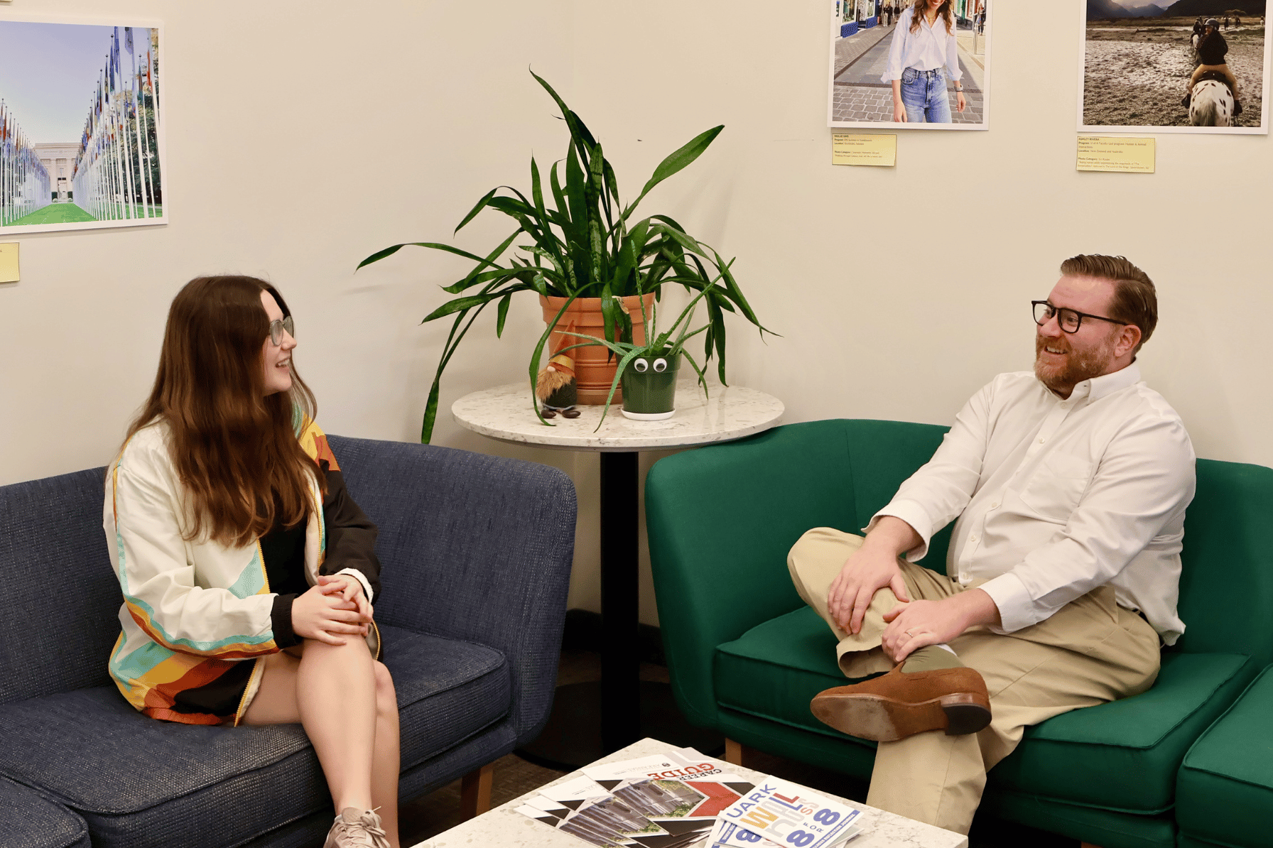 Two people sit on couches having a conversation. There is a table with a plant in between them.