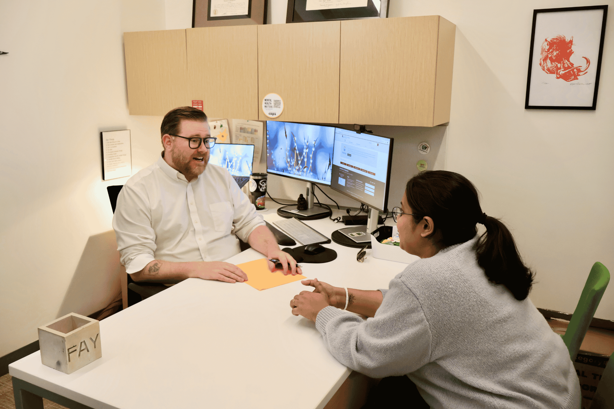 A man and a woman sit at a desk engaged in a conversation. The desk has general office supplies and a computer monitor.