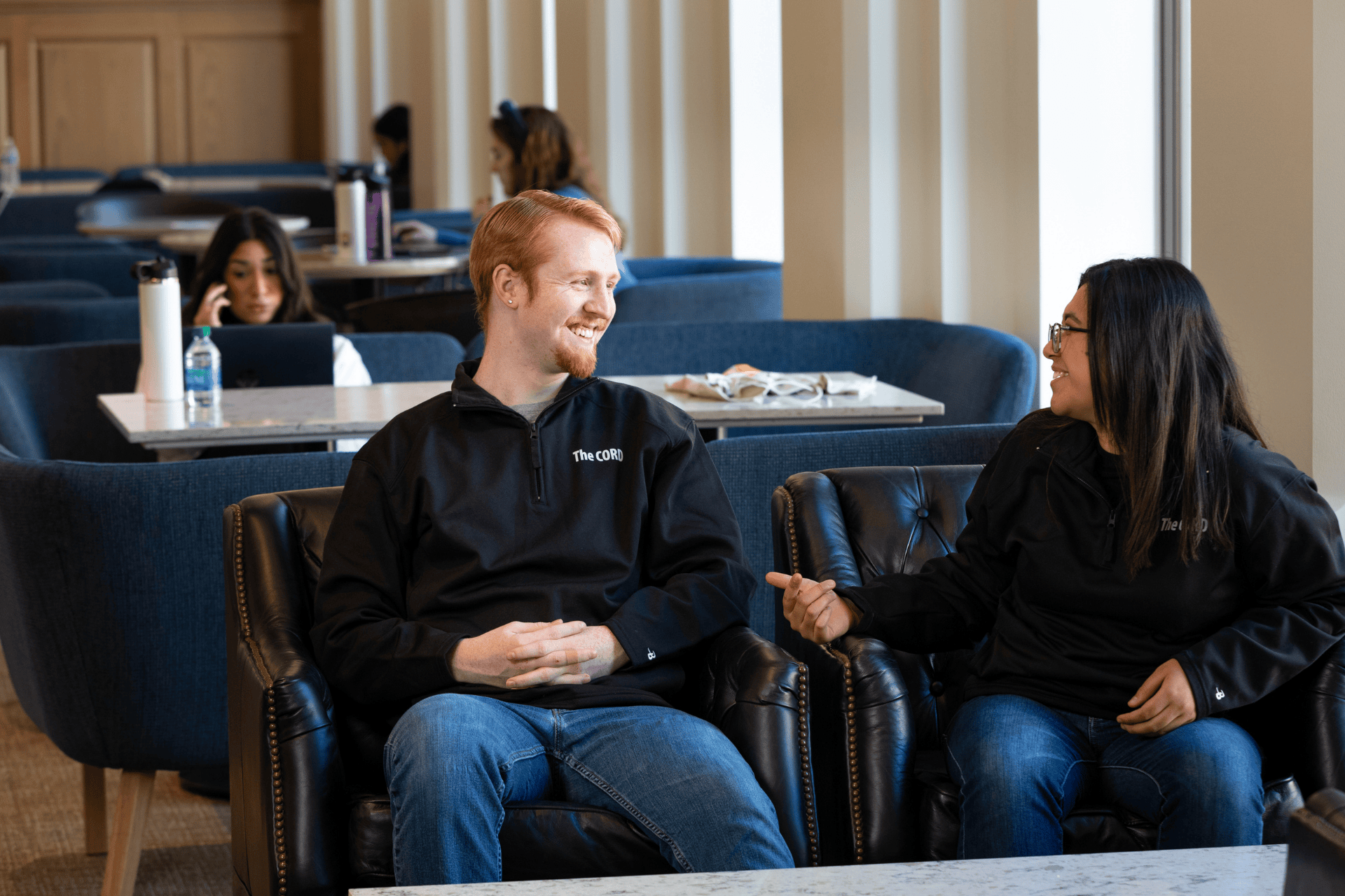 Two students sit on leather chairs and have a conversation. One student has short ginger hair and the other had long dark hair and glasses. They both wear black sweatshirts and blue jeans.